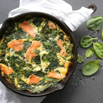 Crustless quiche with salmon and spinach in a cast iron skillet over a white napkin, next to the skillet spinach leaves, salt and pepper scattered over