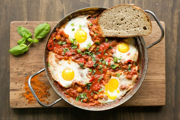 Easy Shakshuka in a large skillet with toasted bread slice on the side, on wood board decorated with spices and basil leaves.