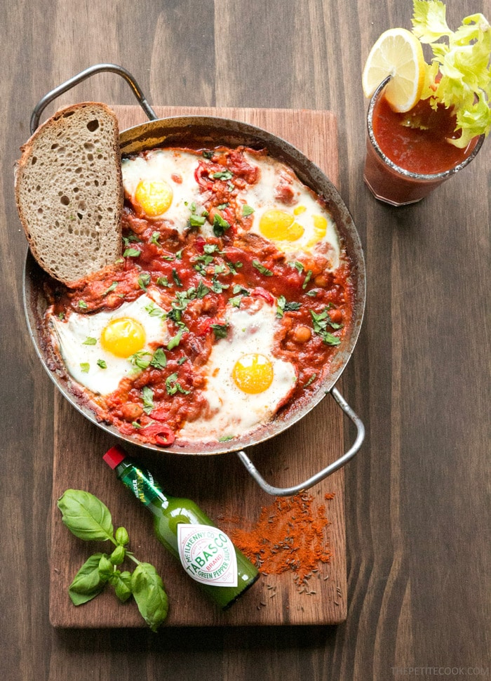 Easy Shakshuka in a large skillet with toasted bread slice on the side, on wood board decorated with spices, green tabasco bottle, and basil leaves. Bloody mary glass on the side.