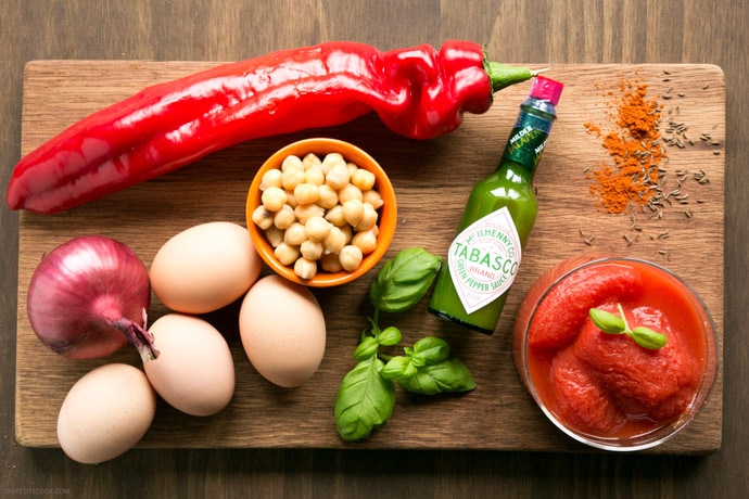 Easy shakshuka ingredients on wood board: one red onion, one long bell pepper, four eggs, a small cup with chickpeas, basil leaves, green tabasco bottle and cup with canned whole tomatoes.