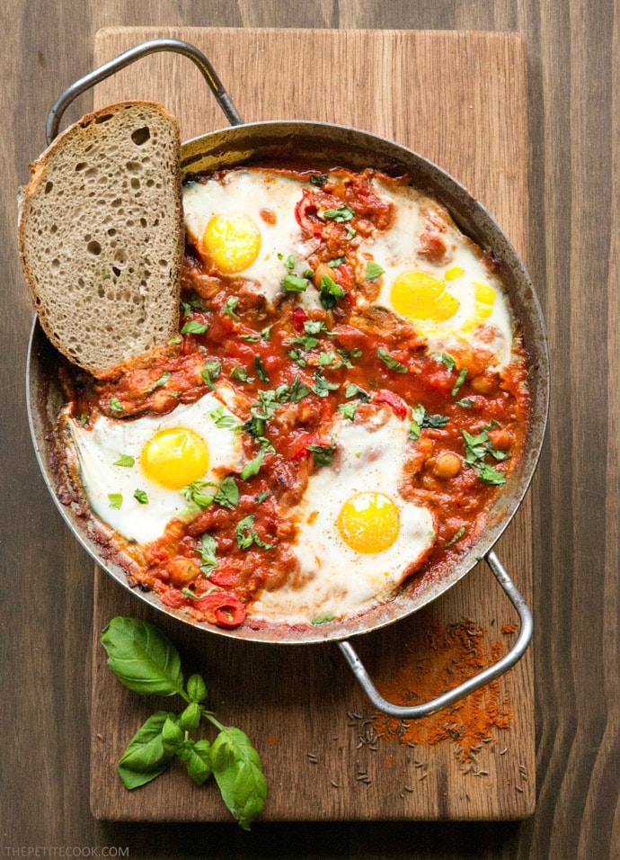 Easy Shakshuka in a large skillet with toasted bread slice on the side, on wood board decorated with spices and basil leaves.