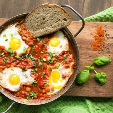 Easy Shakshuka in a large skillet with toasted bread slice on the side, on wood board decorated with spices and basil leaves.