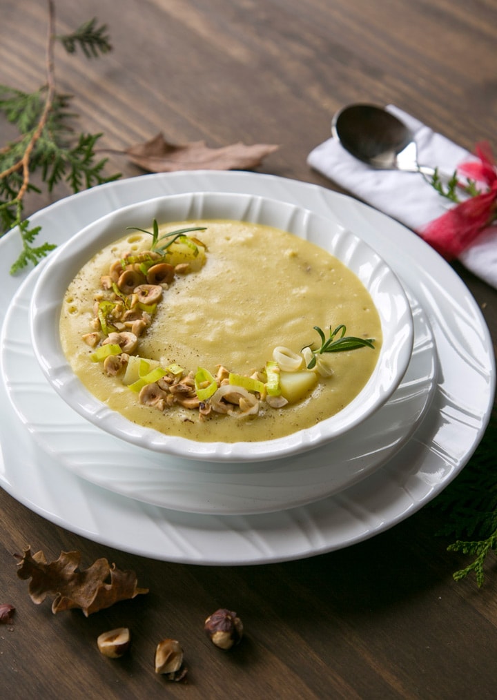 Leek and Potato soup in white bowl over two white plates, spoon on the side over a white napkin. brown and green leaves, and toasted hazelnuts scattered over the wood table