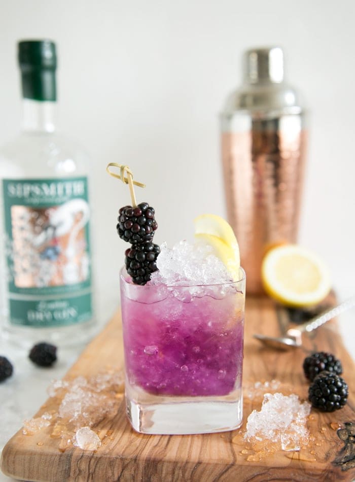 brambe cocktail in a glass topped with 2 blackberries and two lemon slices, over a wood board with crushed ice, blackberries, half lemon, cocktail shaker and gin bottle in the background