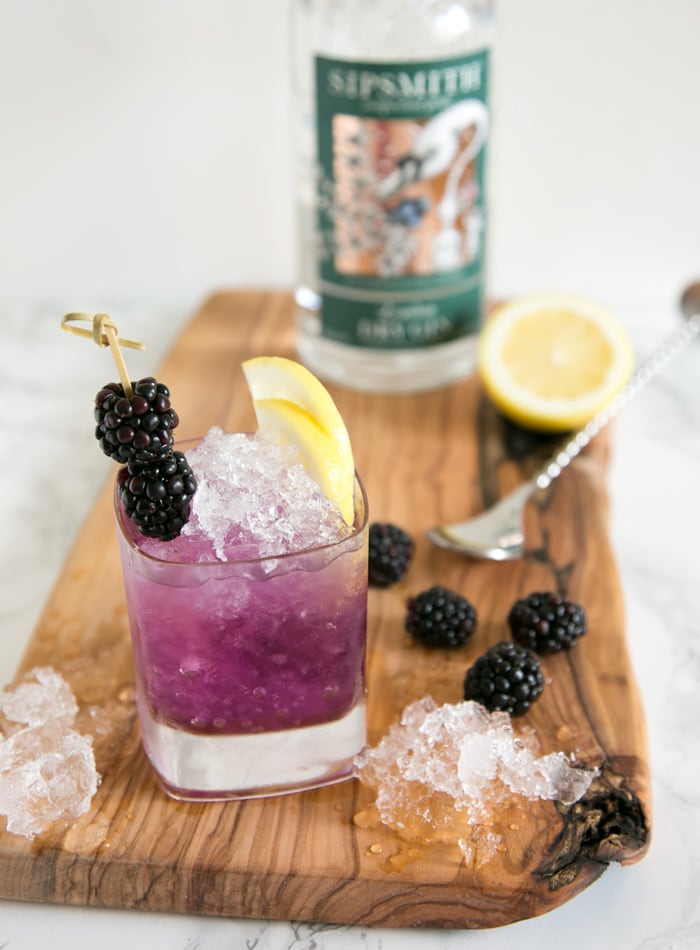 brambe cocktail in a glass topped with 2 blackberries and two lemon slices, over a wood board with crushed ice, blackberries, half lemon, spoon, and gin bottle