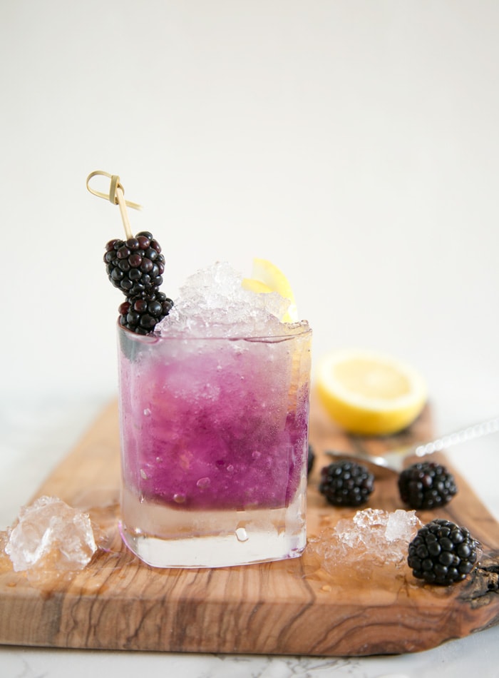 brambe cocktail in a glass topped with 2 blackberries and two lemon slices, over a wood board with crushed ice, blackberries, half lemon. 