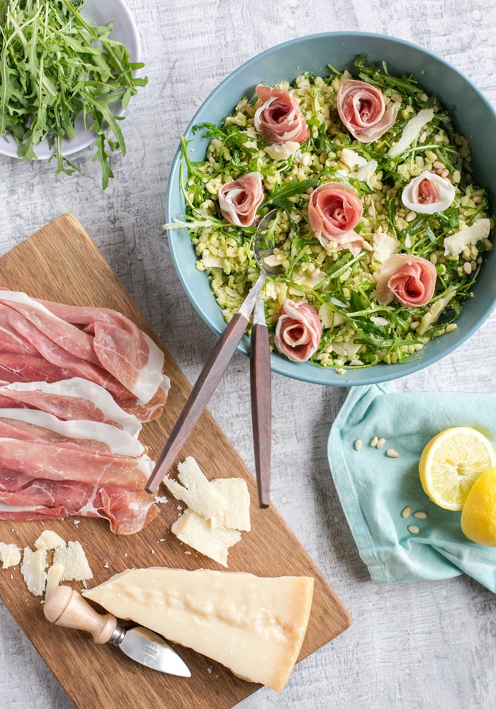  Barley Pea Pesto Salad in a large blue salad bowl with salad spoons. Next to a wood board with prosciutto slices and grana padano cheese piece, and cheese knife. Small bowl of fresh arugula on top, light blue napkin with a halved lemon on the right down side.