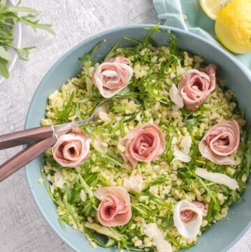 Barley Pea Pesto Salad in a large blue salad bowl with salad spoons. Light blue napkin and halved lemon on the left side, and bowl with arugula on the top left side.