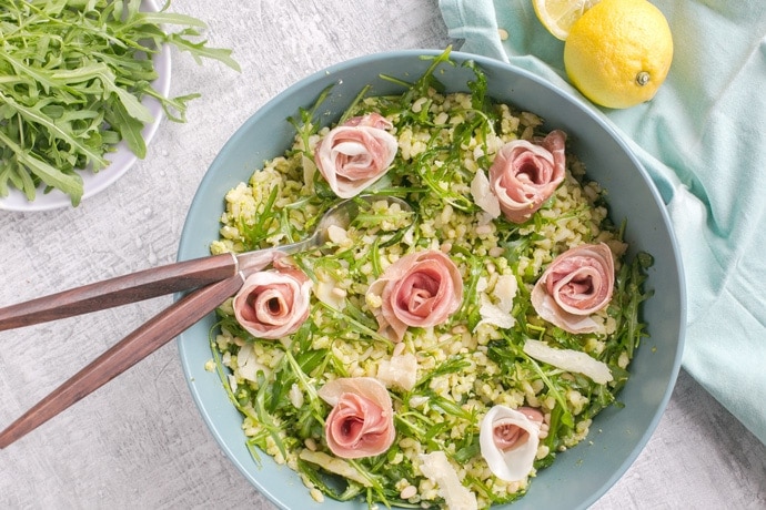 Barley Pea Pesto Salad in a large blue salad bowl with salad spoons. Light blue napkin and halved lemon on the left side, and bowl with arugula on the top left side.
