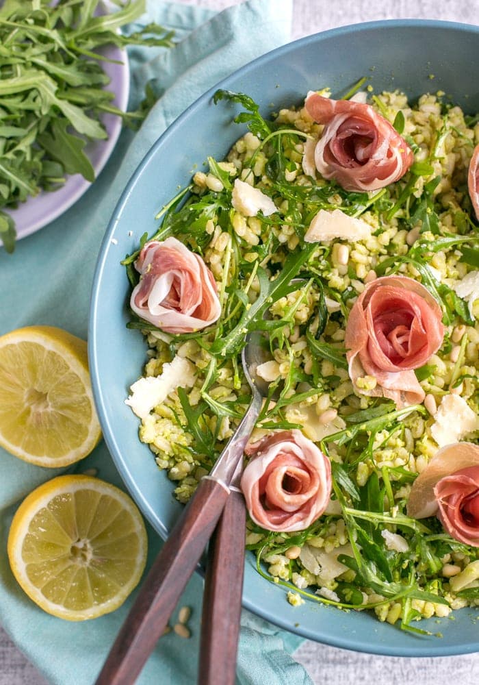  Barley Pea Pesto Salad in a large blue salad bowl with salad spoons. Light blue napkin and halved lemon on the left side, and bowl with arugula on the top left side.