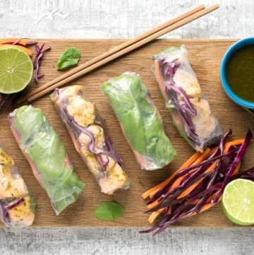 vietnamese summer rolls, chopsticks, halved lime, carrot sticks and sliced cabbage, small blue pot with cilantro dipping sauce on a wood board
