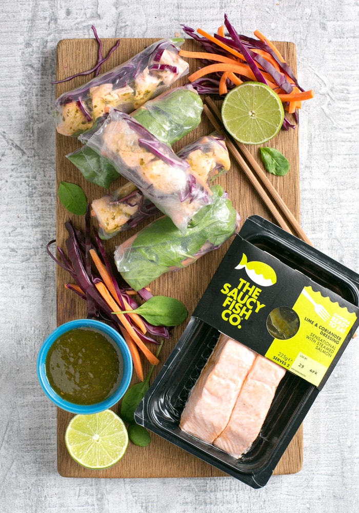 Vietnamese summer rolls on a wood board with halved lime, chopsticks, the saucy fish co salmon fillets packaging and a small white pot with cilantro dipping sauce.