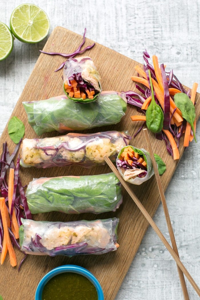 Vietnamese summer rolls on a wood board with halved lime, chopsticks and a small blue pot with cilantro dipping sauce.