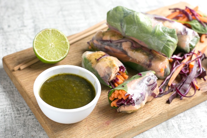 vietnamese summer rolls on a wood board with halved lime, chopsticks and a small white pot with cilantro dipping sauce.
