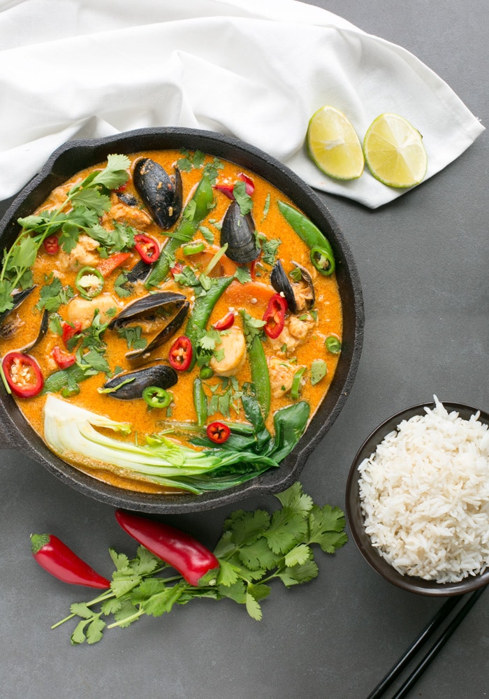 Thai Red Fish Curry in cast iron skillet, next to fresh cilantro, red chili peppers and halved limes, a small bowl with rice and black chopsticks.