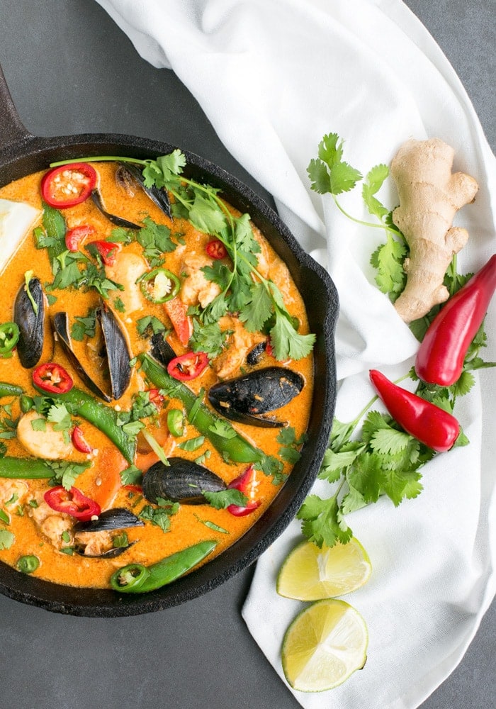 Thai Red Fish Curry in cast iron skillet, next to a white cloth with fresh ginger root, cilantro, red chili peppers and halved limes. 