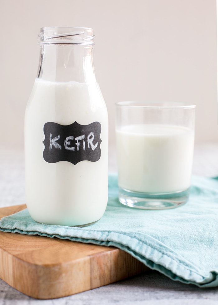 Homemade kefir into small glass bottle, glass with kefir next to it over a light blue napkin and wood board