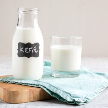 Homemade kefir in small glass bottle next to a glass with kefir, over light blue napkin and wood board