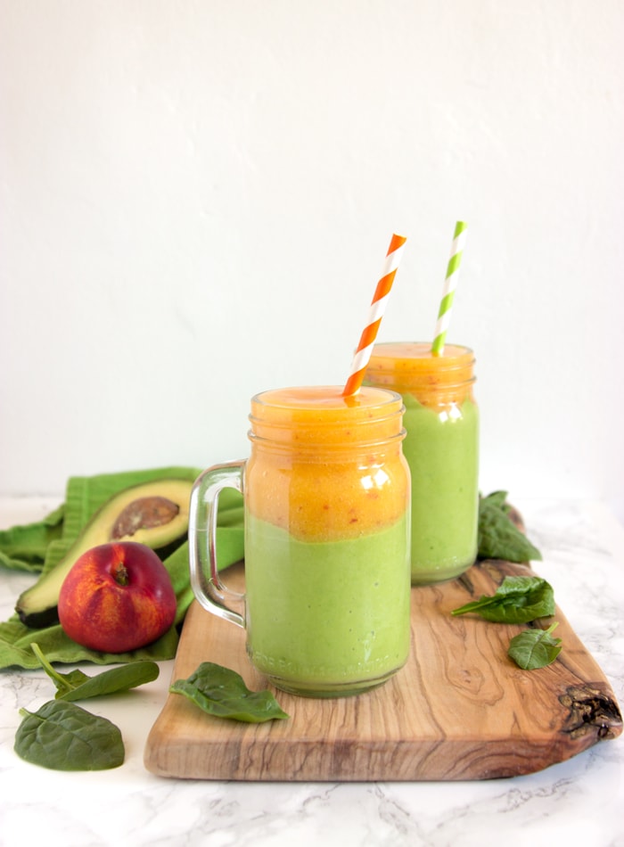 2 drinking jar filled with nectarine and avocado smoothie on a wood board, next to a nectarine, spinach leaves and halved avocado.