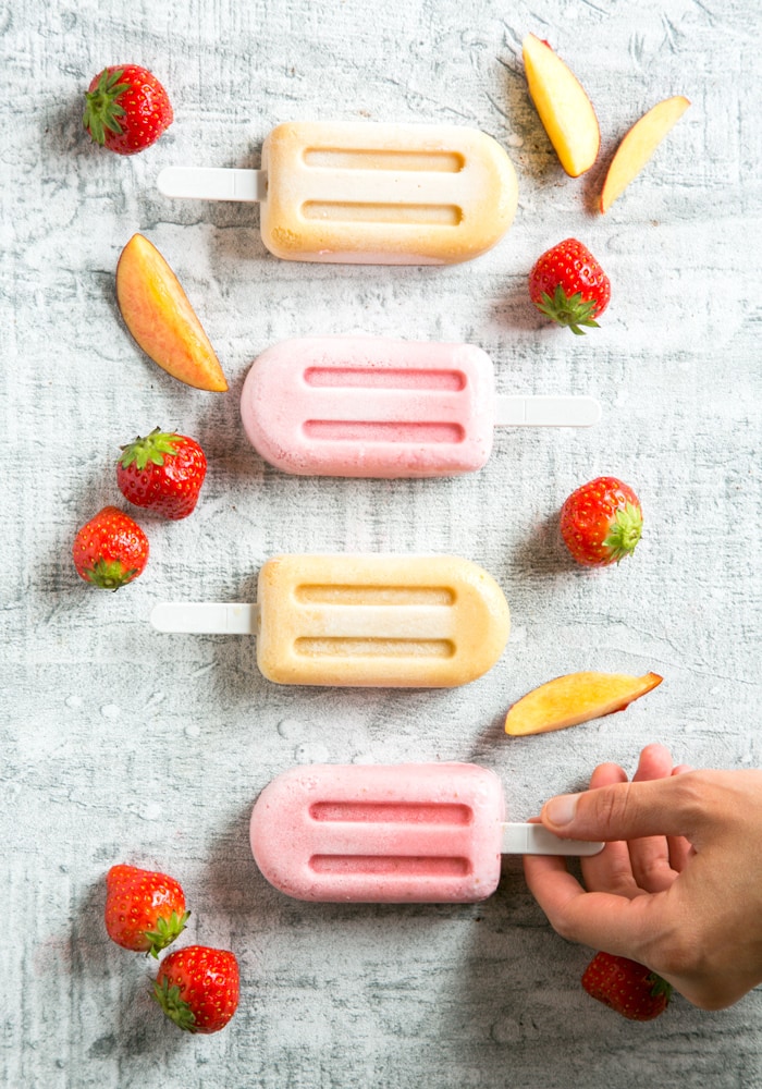  Nectarine and Strawberry Smoothie Popsicles on grey background, hand holding one popsicles stick. strawberries and nectarine slices scattered around