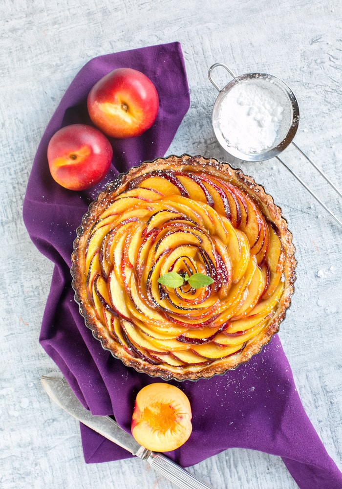 nectarine tart over a purple napkin, two nectarines on the top left, a sieve with powdered sugar on the top right side, half nectarine and a knife on the bottom, 