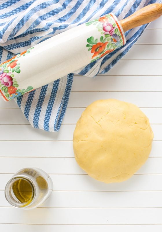 olive oil pastry dough next to a glass of olive oil, on the top side a white rolling pin over a blue and white striped napkin