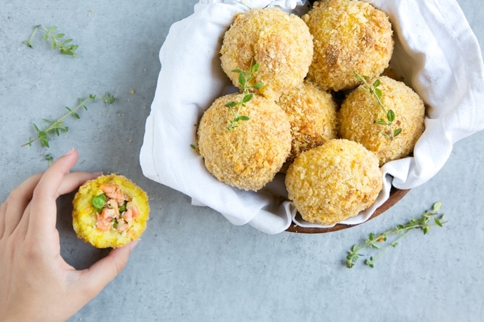 salmon and pea baked arancini rice balls in a dish covered with white napkin, on the left side hand holding half arancini ball
