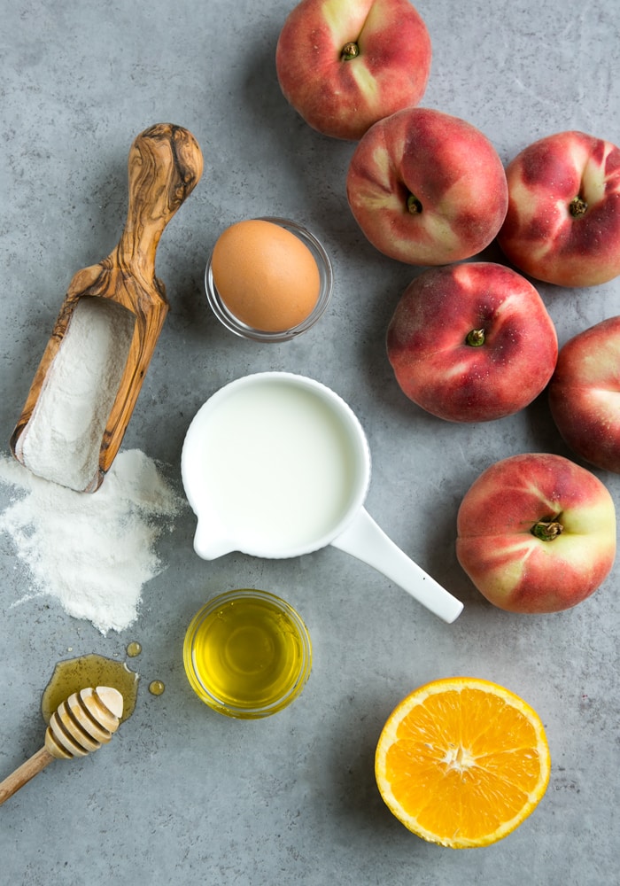 flour, egg, peaches, olive oil, honey, half orange and buttermilk in a cup on grey background