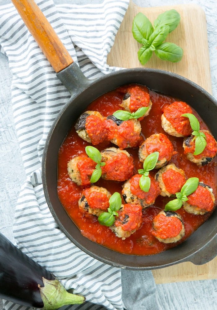  Vegetarian Aubergine Meatballs in pan over striped napkin and wood chopping board, aubergine on the left-down corner and basil leaves on the top right corner.