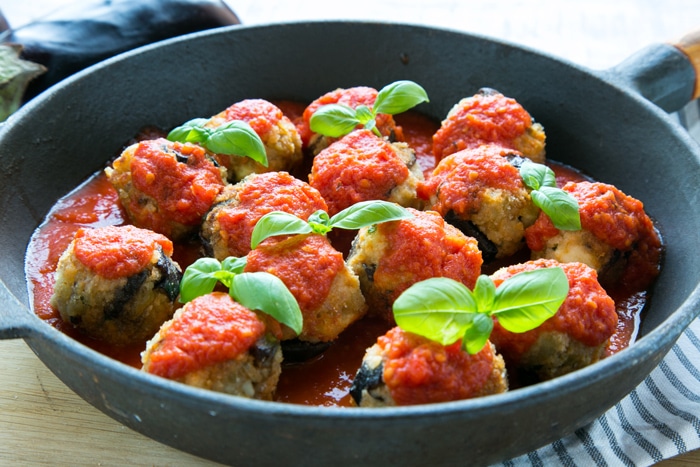 Vegetarian Aubergine Meatballs topped with tomato sauce and basil leaves in a large cast iron skillet over striped napkin, side view.