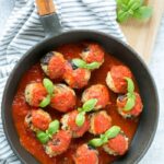 eggplant meatballs with tomato sauce and basil leaves in a cast iron skillet, white and grey stiped napkin beneath and a wood board.