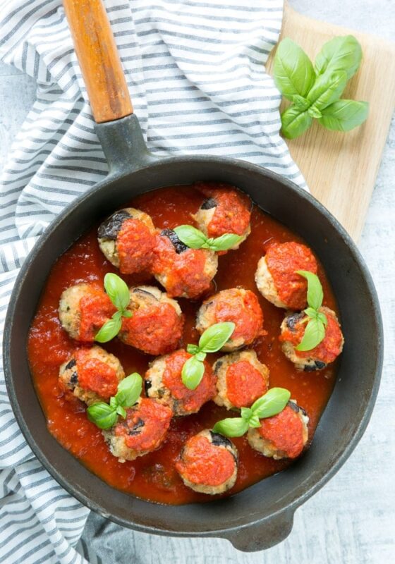 vegetarian eggplant meatballs with tomato sauce and basil leaves in a cast iron skillet, white and grey stiped napkin beneath and a wood board.