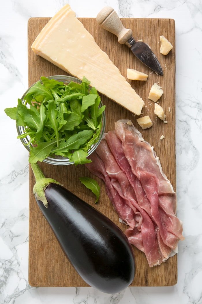 Eggplant, prosciutto, grana padano cheese, cheese knife and rocket leaves in glass bowl all placed over a wood board