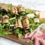 eggplant roll-ups on a bed of rocket leaves next to prosciutto slices and grana padano cheese chunks all placed on a wood board