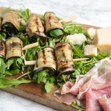 eggplant roll-ups on a bed of rocket leaves next to prosciutto slices and grana padano cheese chunks all placed on a wood board