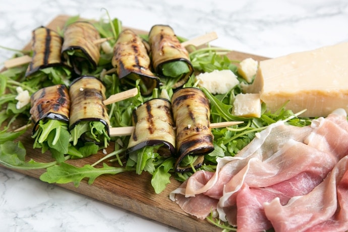 eggplant roll-ups on a bed of rocket leaves next to prosciutto slices and grana padano cheese chunks all placed on a wood board