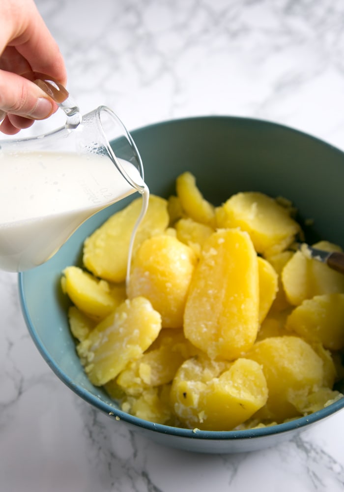 recipe step 2: hand pouring hot milk into the bowl with lightly mashed potatoes with butter