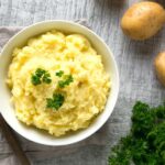 best mashed potatoes topped with parsley in a white bowl over a grey napkin, potatoes and parsley in the background
