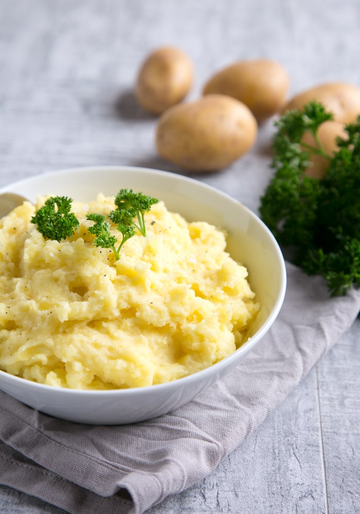 the best mashed potatoes, topped with parsley in a white bowl over a grey napkin, potatoes and parsley in the background