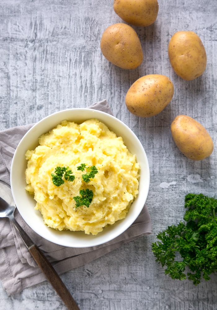 mashed potatoes topped with parsley in a white bowl over a grey napkin and serving spoon, potatoes and parsley next to the bowl