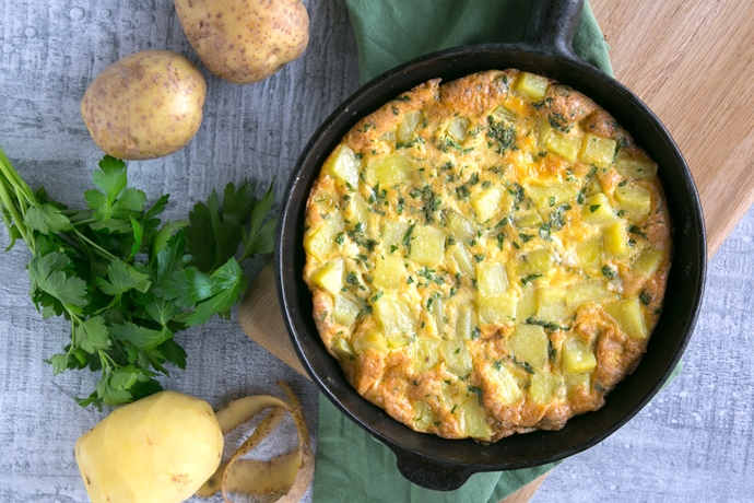 recipe step 3: Italian Potato Frittata in a cast iron skillet over a wood board with green napkin, potatoes and parsley on the background.