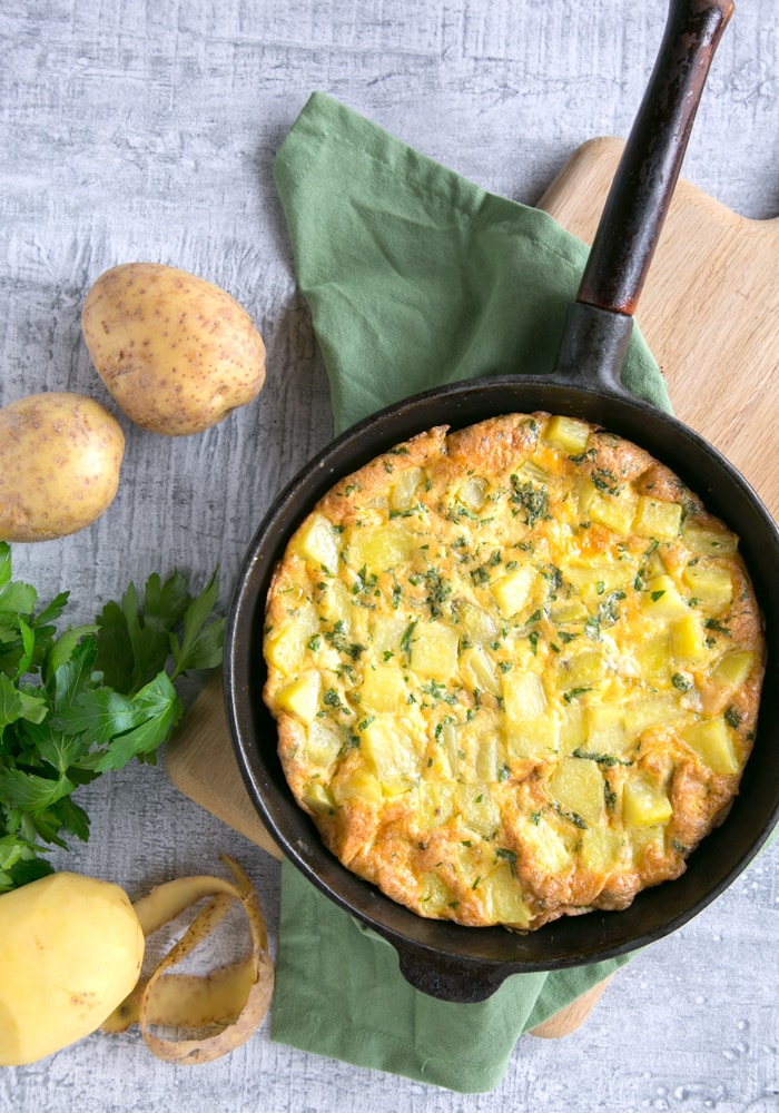 Classic Italian Potato Frittata in a cast iron skillet over a wood board with green napkin, potatoes and parsley on the background.