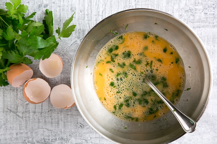 recipe step 2: whisked eggs and parsley in a bowl, next to egg shells and parsley leaves