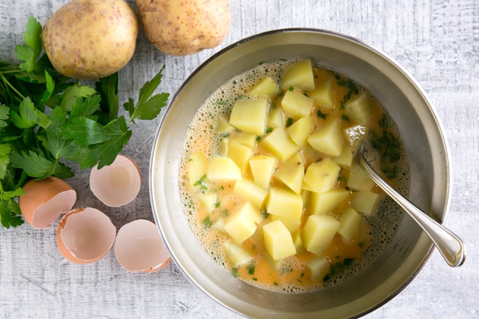 whisked eggs, parsley and chopped potatoes in a large bowl, next to egg shells parsley and potatoes