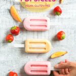four nectarine and strawberry smoothie popsicles on a grey background, one hand holding the stick of one popsicle. Image for Pinterest