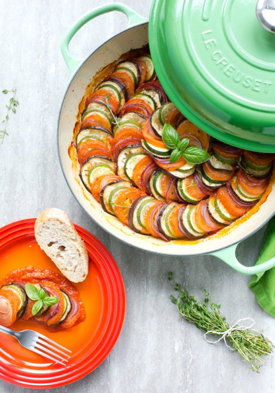 Easy Ratatouille in a large green cast iron shallow pan, bunch of thyme sprigs on the left side and a green napkin on the top right side. Red small dish with a slice of bread a small portion of ratatouille and fork on the left side of the image.