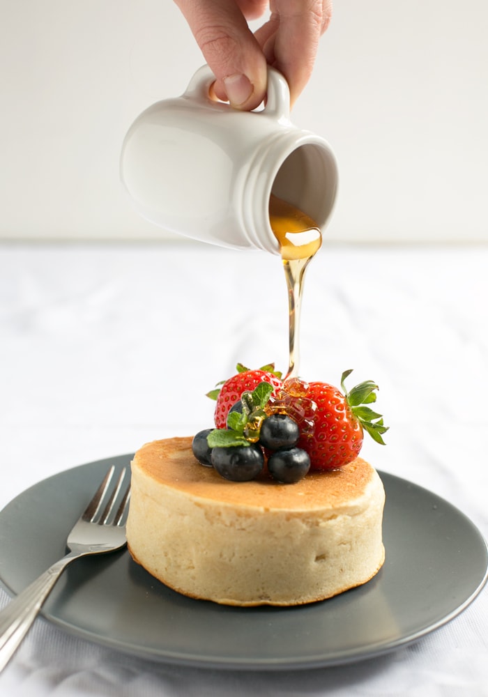 Hand holding small white jar pouring maple syrup on top of Japanese pancakes topped with strawberries and blueberries and fork next to it, on grey plate and white tablecloth