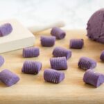 Purple homemade potato gnocchi on a wood board, next to the dough and gnocchi wood board.