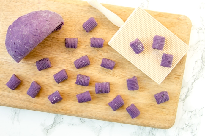 Homemade purple potato gnocchi on a wood board, next to the dough and gnocchi wood board.