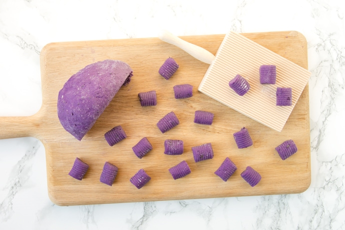 Homemade purple potato gnocchi on a wood board, next to the dough and gnocchi wood board.